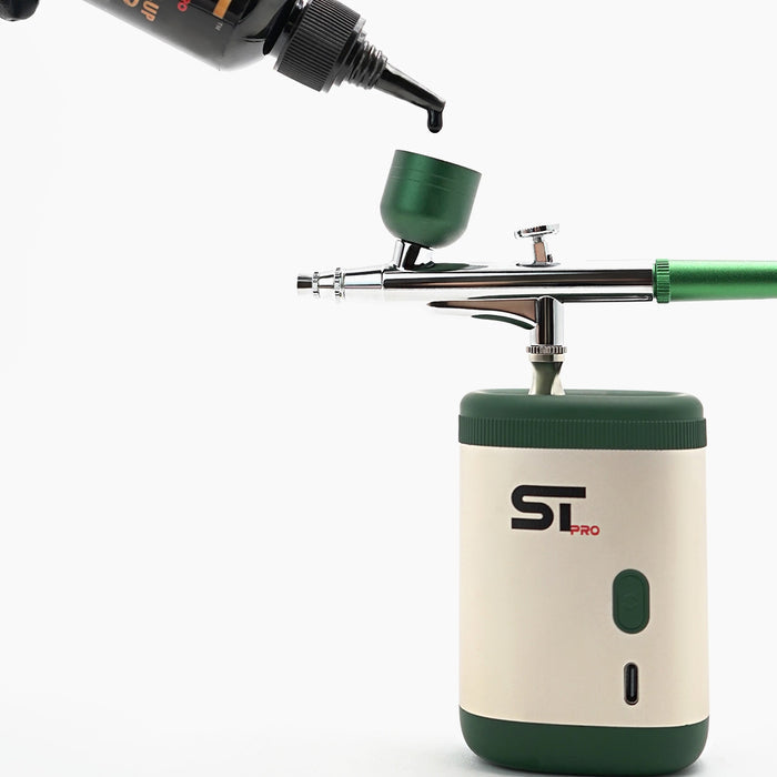 An ST Pro airbrush with a cream and dark green body, shown with a green ink cup and a black bottle of ST Color Liner being dispensed above it on a white background.