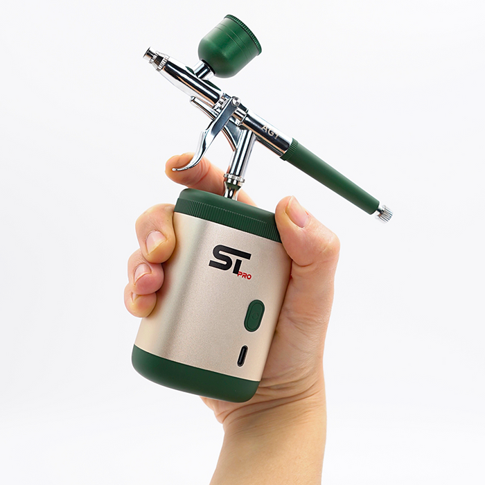 A hand holding an ST2000 airbrush tool with a metallic silver body, dark green accents, and a green cap, set against a plain white background.