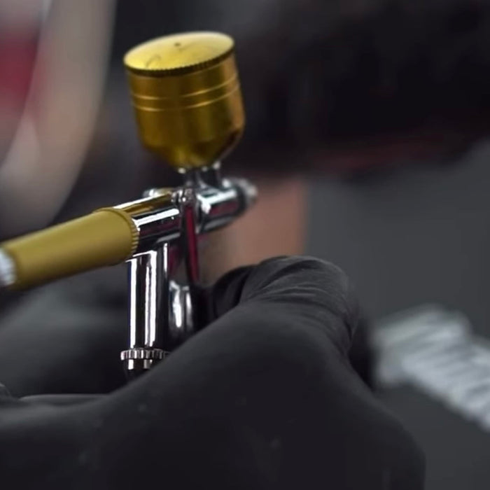 A close-up of an airbrush paint gun with a gold cap and metallic body, held by a hand wearing a black glove. The background is heavily blurred, showing what appears to be a person's head and hair.